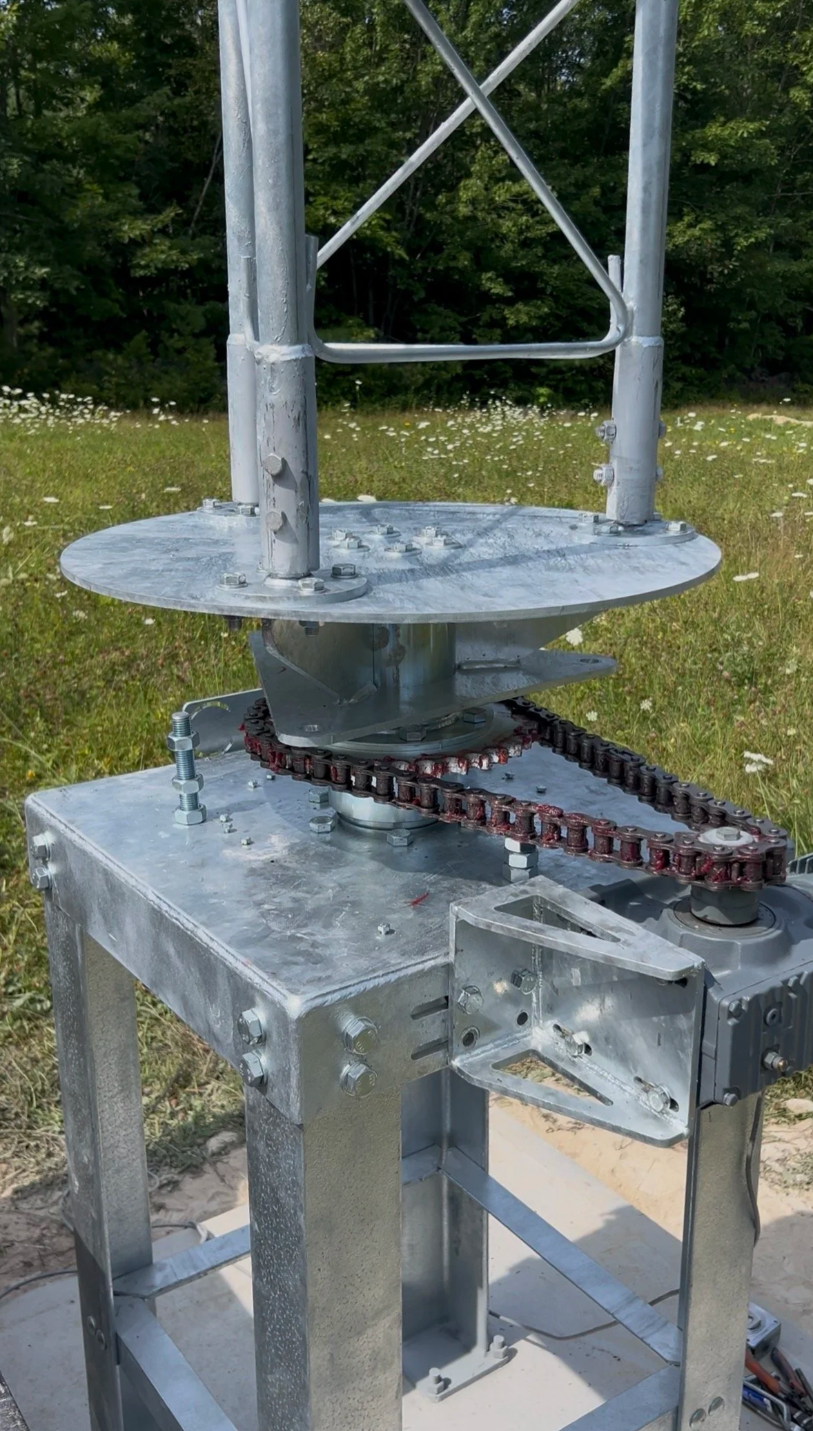 Close-up of a metallic structure with a chain drive system, set outdoors on a grassy area with trees in the background.