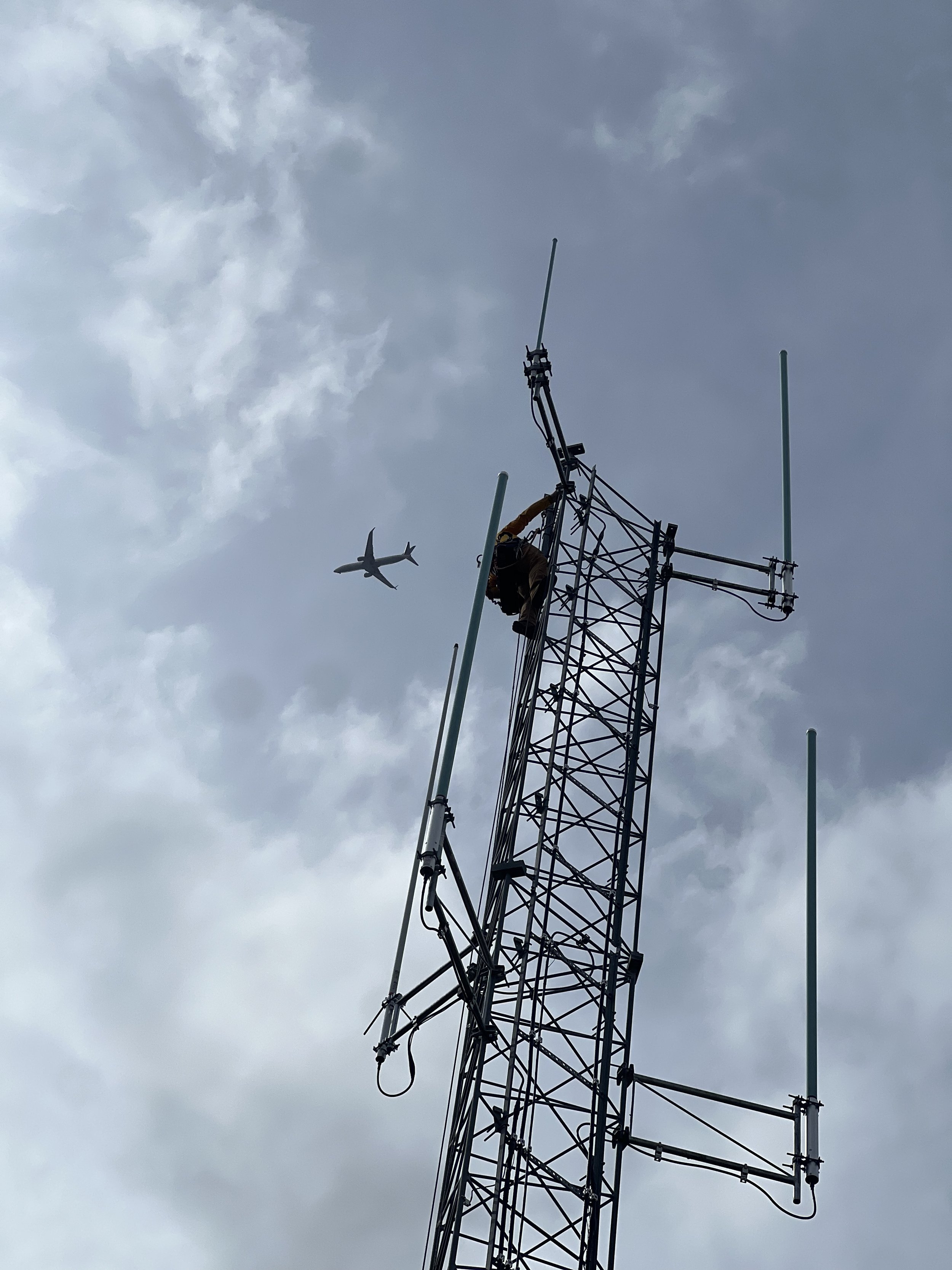 Tower climber performing antenna work with aircraft in background — aviation and communications infrastructure contractor, sterile area qualified