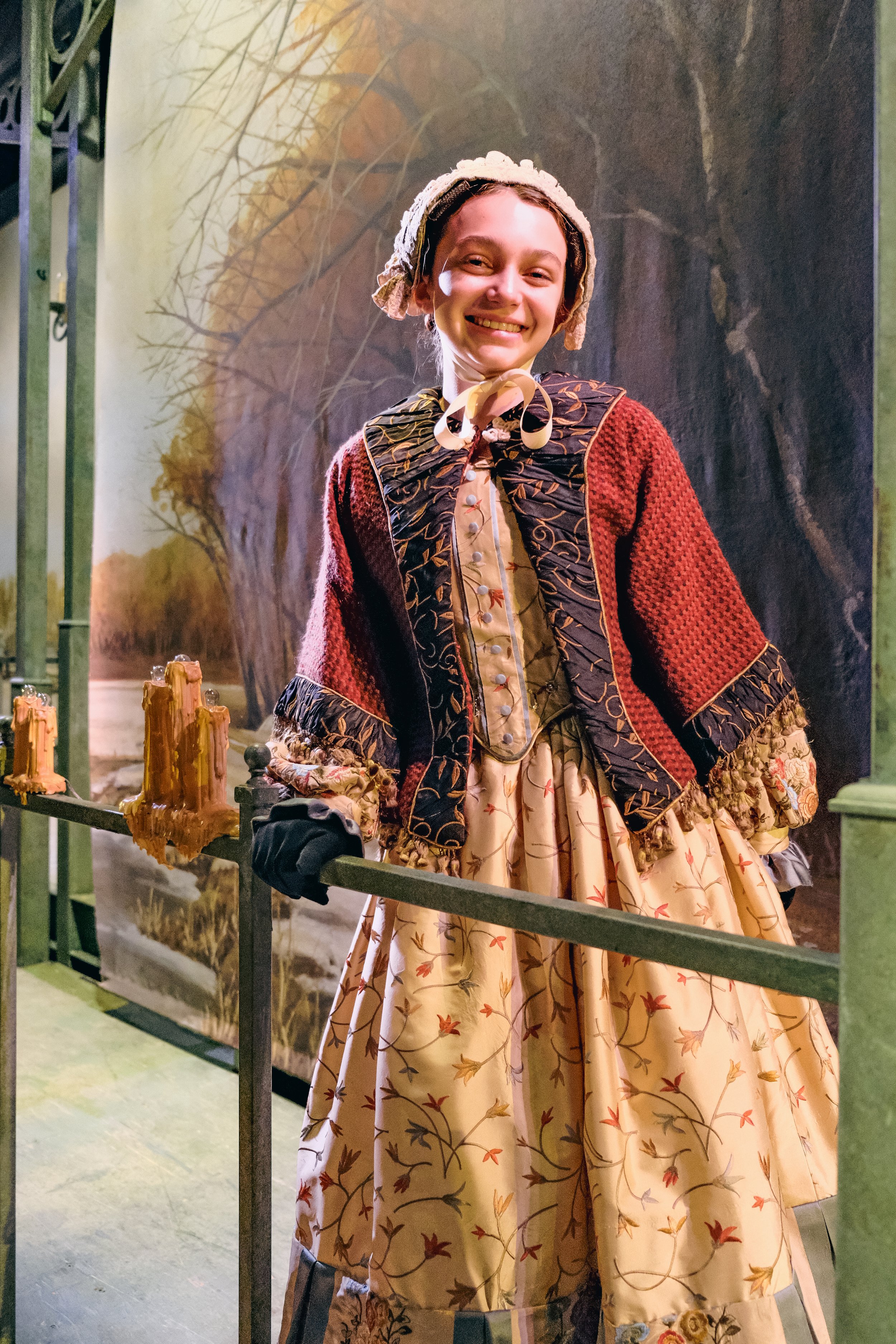 A woman dressed in historical clothing, including a floral patterned dress, a embroidered jacket, and a bonnet, standing on a stage with a painted backdrop of trees and a river, smiling at the camera.