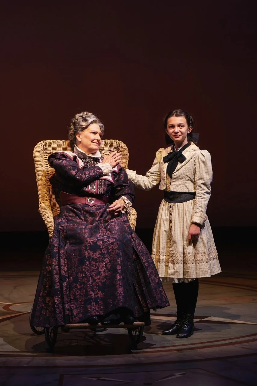 A woman in an antique purple and black dress with lace details seated in a wicker wheelchair, and a young girl in a beige dress with a black ribbon, standing beside her touching her shoulder, on a stage with a dark background.