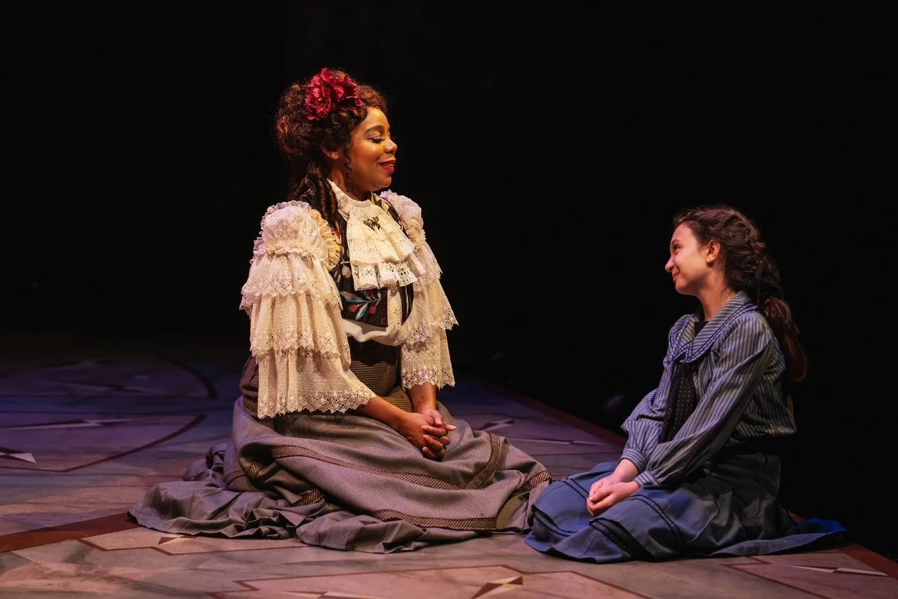 Two women sitting on the stage floor, engaged in a conversation. The woman on the left has curly hair with a red flower accessory and wears a vintage-style lace blouse and a long skirt. The woman on the right has dark wavy hair tied back and wears a striped dress. The background is dark, and the scene is lit with stage lighting.
