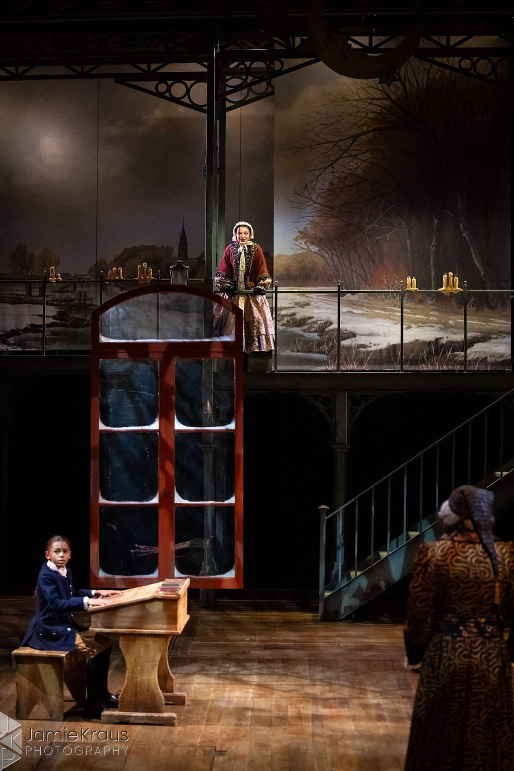 The scene appears to be a theatrical stage set with actors in period costumes. The background features a scenic painting of a river landscape with trees and overcast weather. A woman in 18th-century attire is standing behind a railing, and a young girl, also in period clothing, is sitting at a wooden desk with a book in a classroom setting. An adult woman in traditional clothing is observing the scene from the right side. The overall atmosphere suggests a historical or classical play.