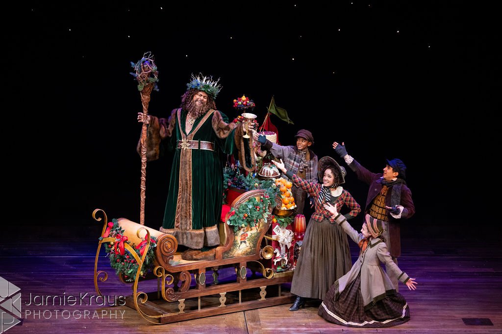 Actors dressed in costumes perform on stage with a large Christmas-themed sleigh filled with presents and decorations, celebrating a festive holiday scene.
