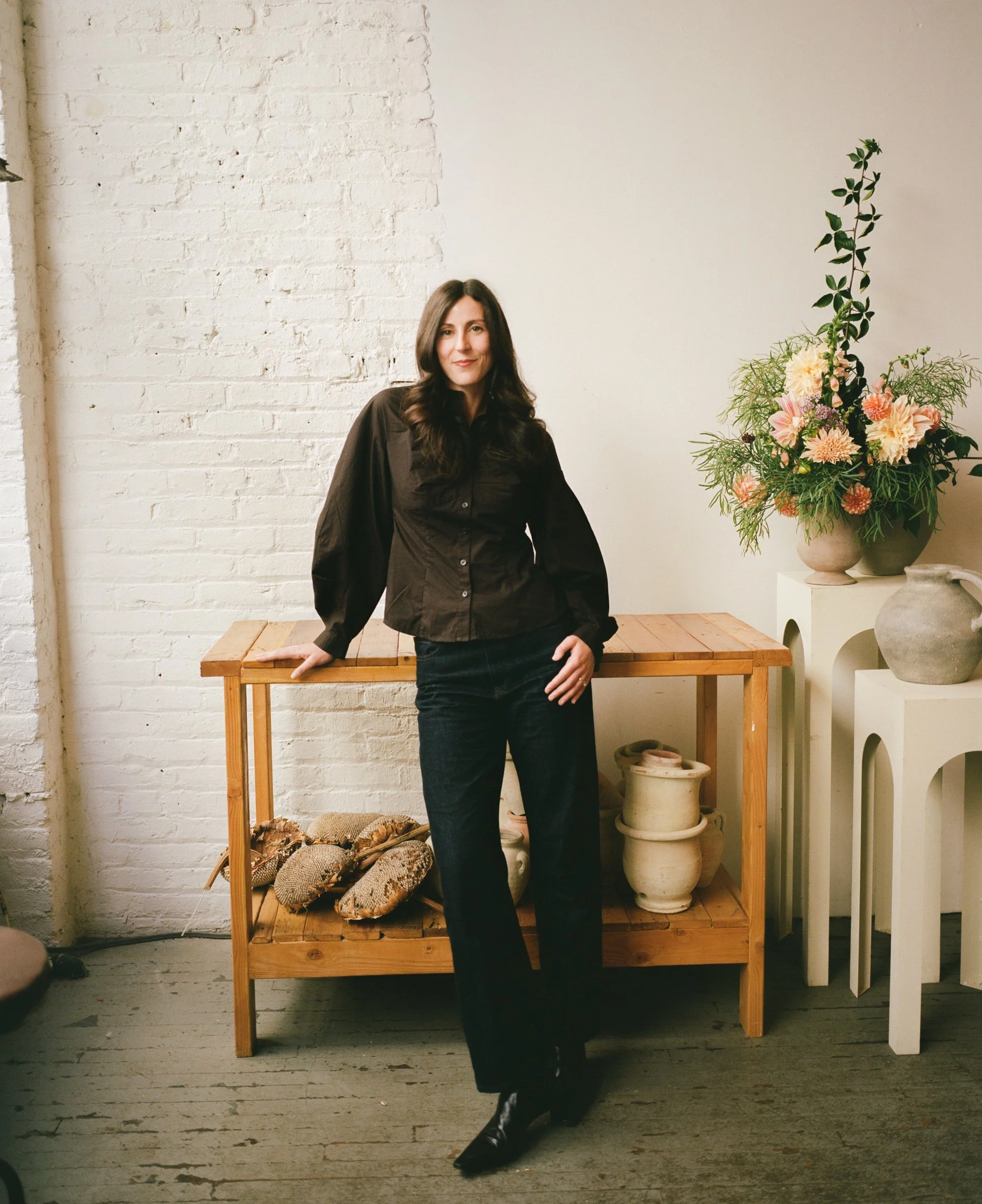 A woman with long dark hair leaning against a wooden table in a room with white brick wall and minimal decor, including a large floral arrangement and pottery.