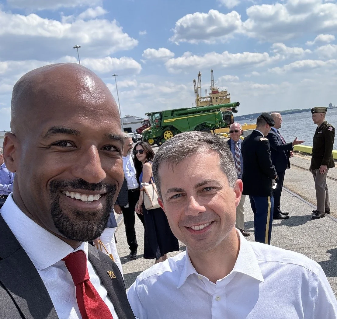 Mark taking a selfie with Pete Buttigieg, U.S. Secretary of Transportation. Several people in the background, some military personnel, and a green farm equipment vehicle on the dock.