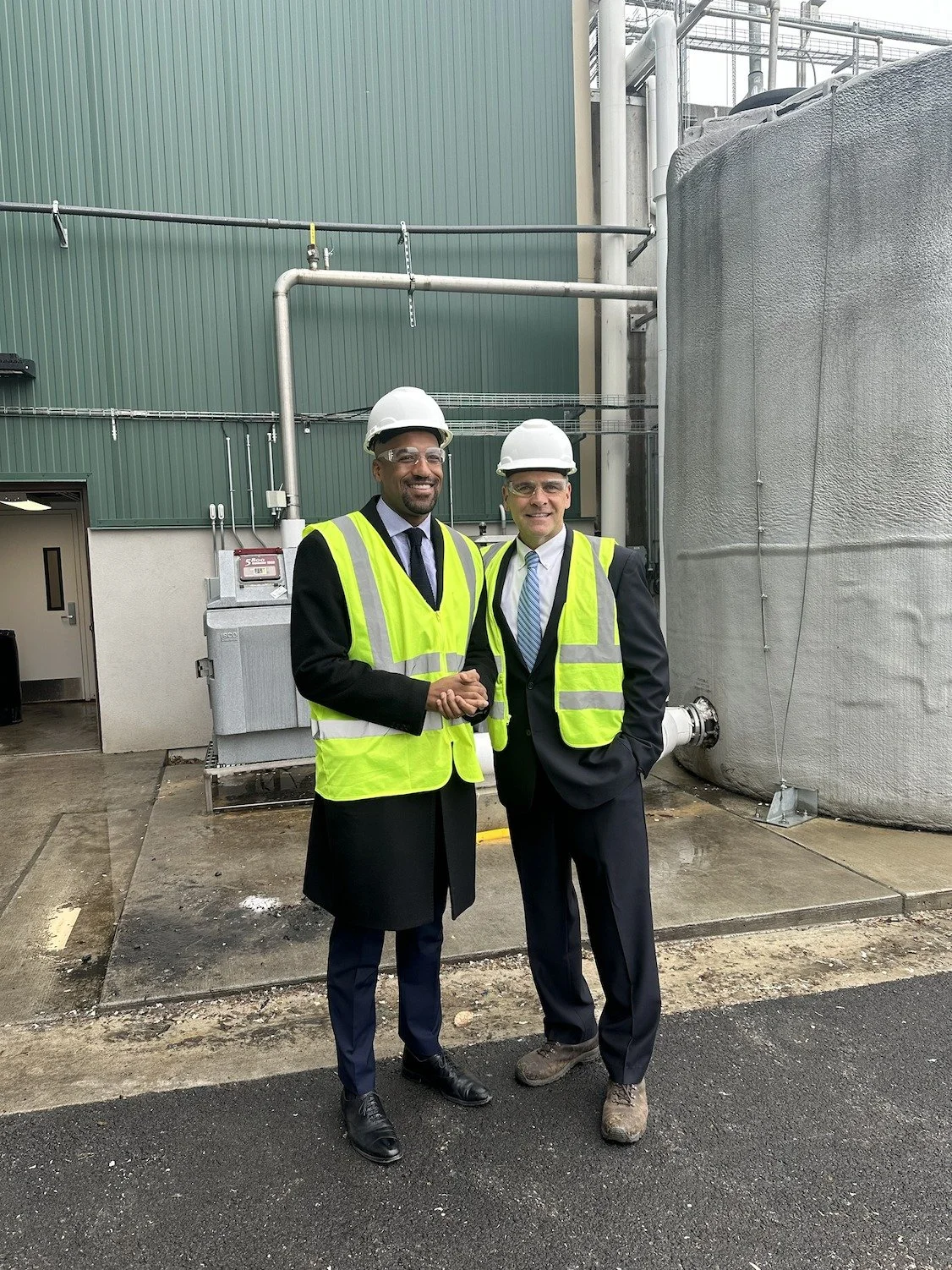 Mark and another man in business suits and safety vests shaking hands outdoors at an industrial site, both wearing white hard hats.