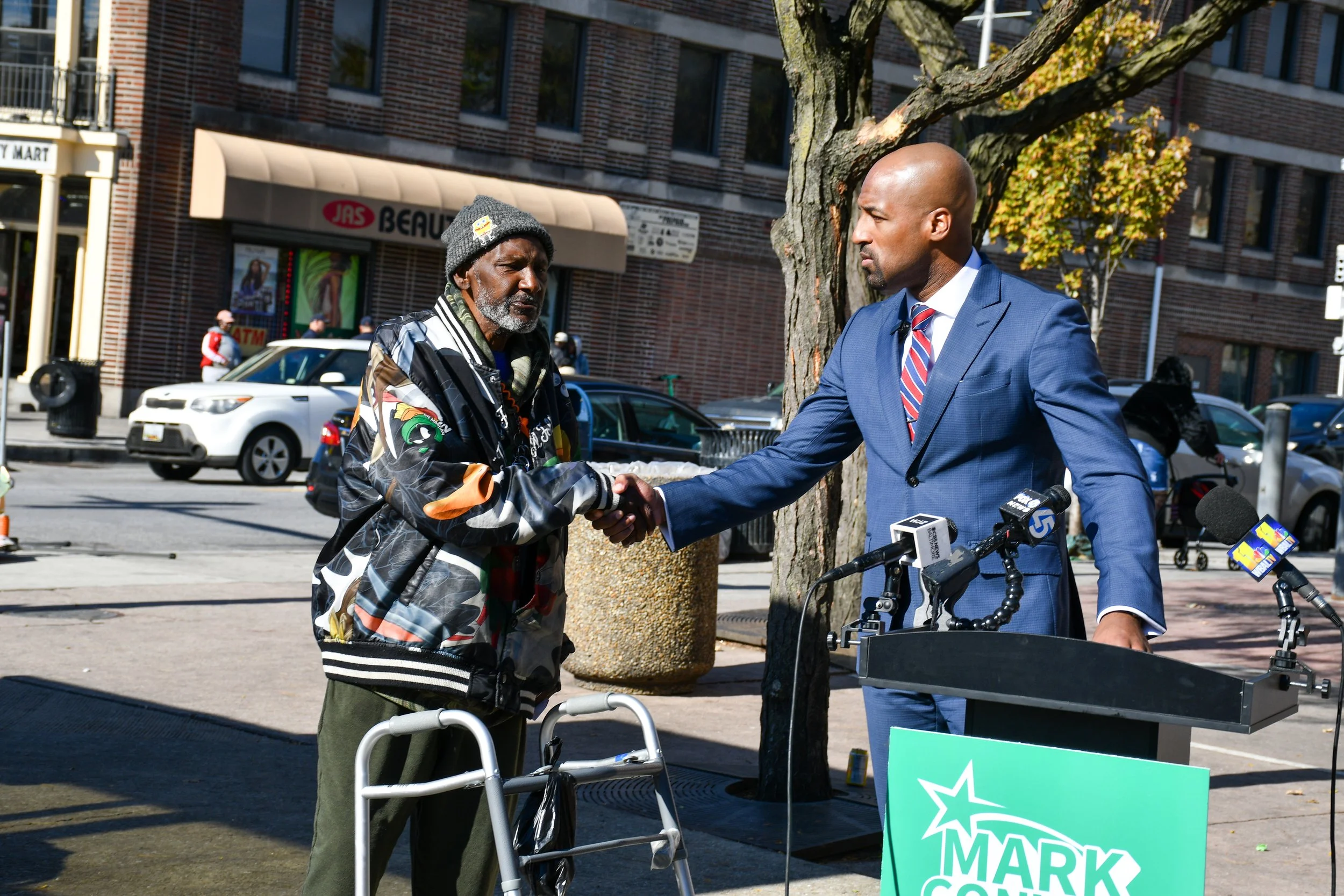 Mark in a suit standing behind a podium, shaking hands with a man with a walker on a city street during a press event.
