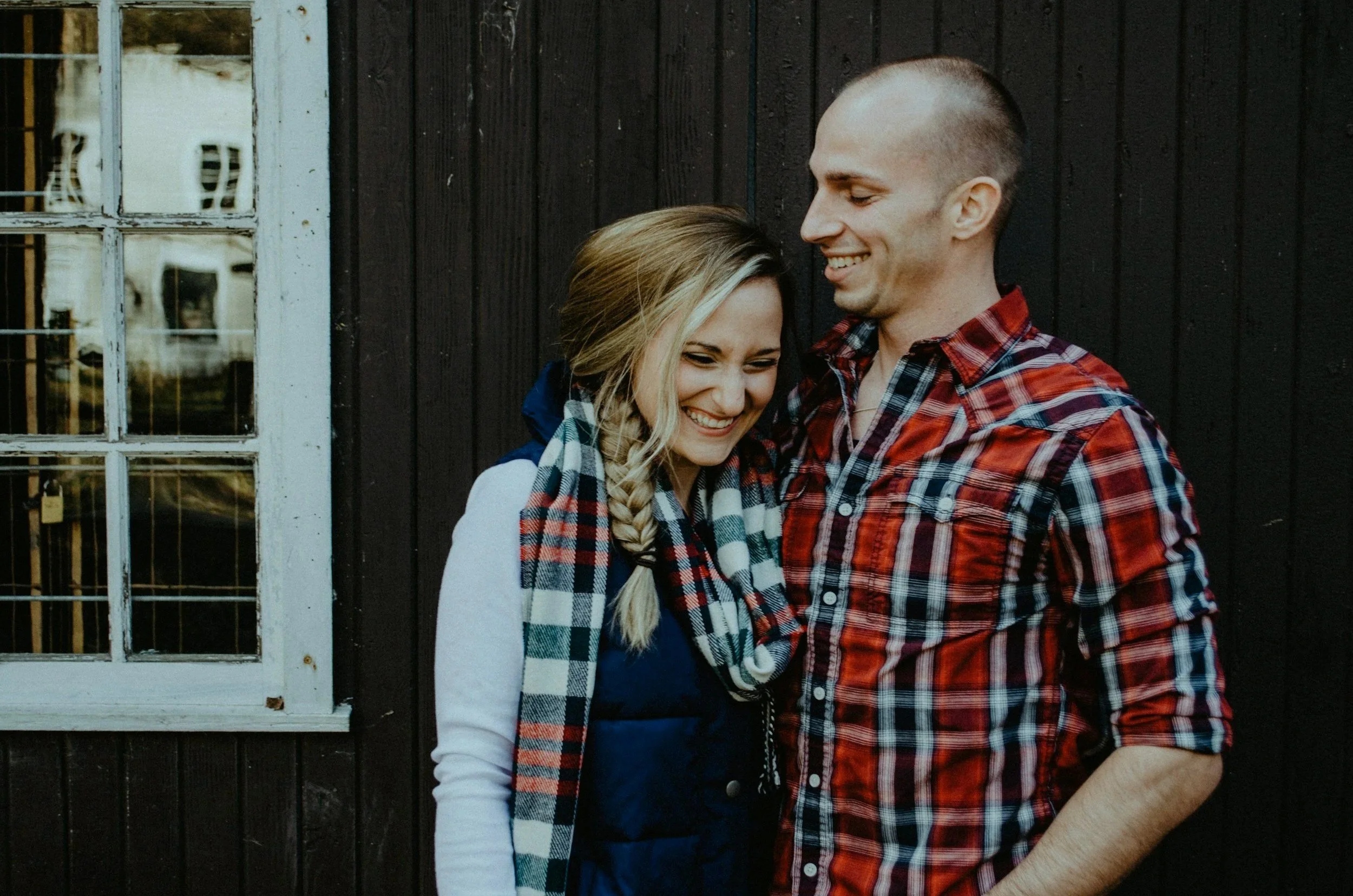 A smiling man and woman, who've been in therapy, standing close together in front of a dark wood wall, with the woman wearing a checkered scarf and the man wearing a red plaid shirt.