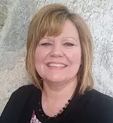 A woman with blonde hair smiling, wearing a black top and a necklace, standing outside in front of a stone wall.