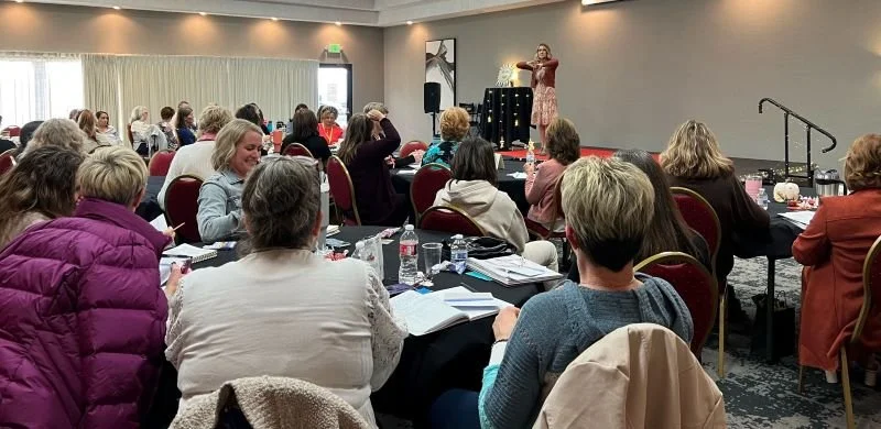 Women attending a conference in a ballroom, listening to a speaker on stage.