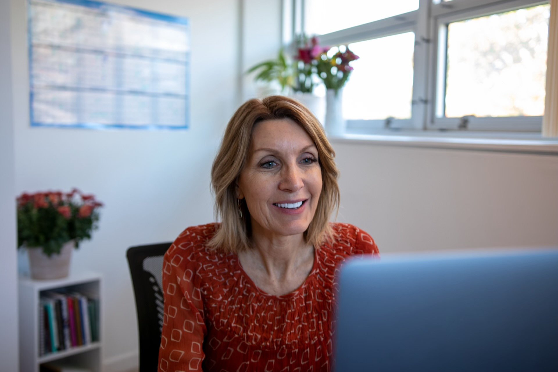 Carolyn Cooper with shoulder-length light brown hair smiling at a laptop in an office with white walls, a window, a group of pink flowers in a white pot, and a small bookshelf.