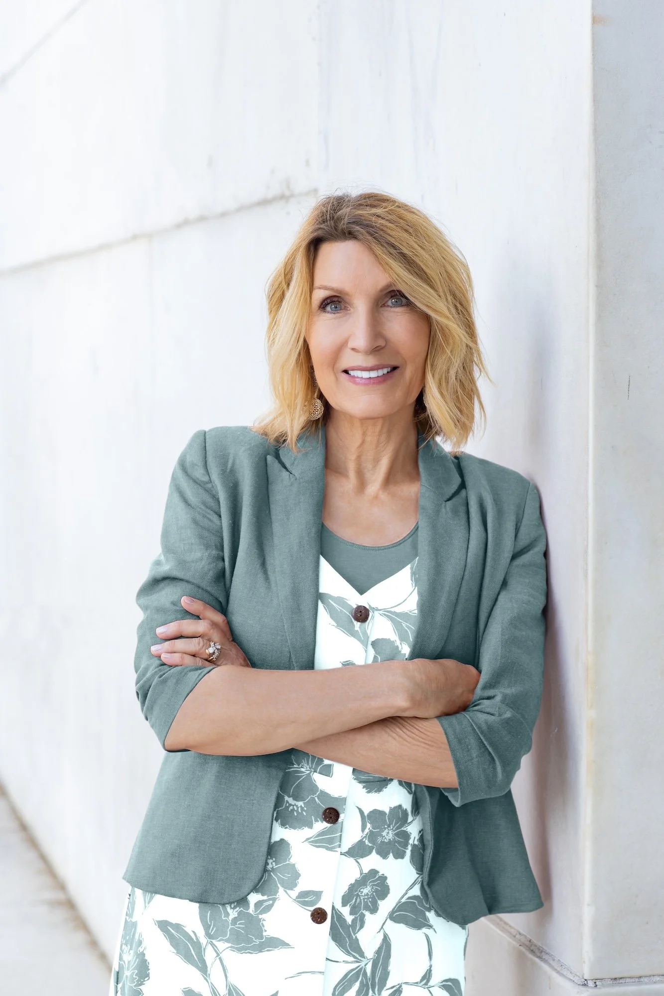 A middle-aged woman with blonde hair and blue eyes, wearing a green blazer over a floral dress, stands outdoors against a light-colored concrete wall, smiling with arms crossed.