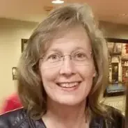 A woman with shoulder-length light brown hair and glasses, smiling indoors in a softly lit room.