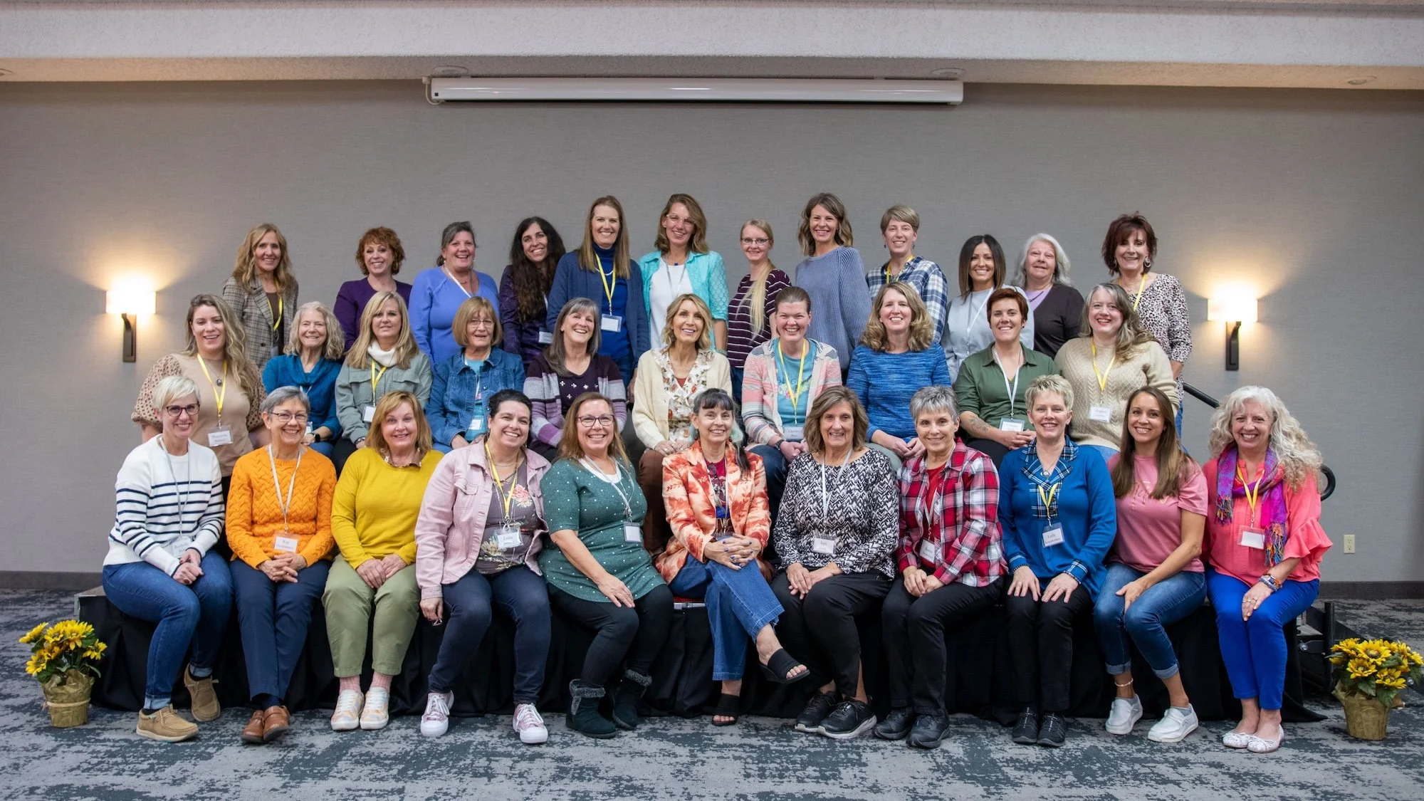 Group of diverse women posing for a photo on stage at a conference or event, with a plain gray wall and sconce lights in the background
