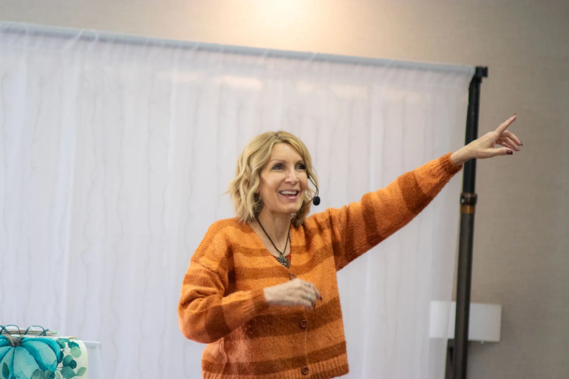 A woman with shoulder-length blonde hair, wearing an orange and brown striped sweater and a microphone headset, is smiling and pointing to her right in an indoor setting with a beige and gray wall background.