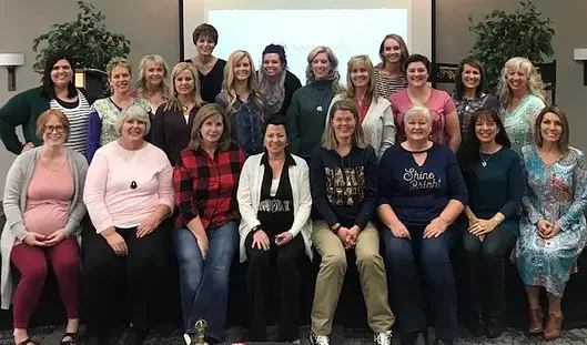 Group of women gathered in a room for a photo, some sitting and others standing in the background, with a screen behind them, indicating a possible seminar or meeting.