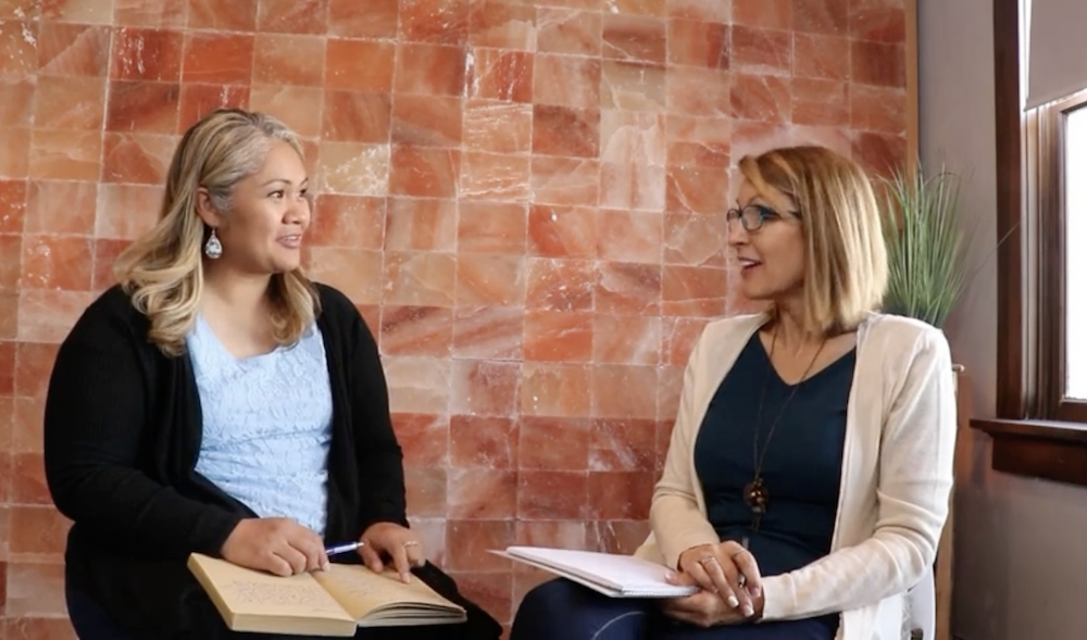 Two women sitting and talking in a room with a salt-colored tiled wall. One woman has blonde hair, wearing a light blue top and black cardigan, holding an open book and a pen. The other woman has red hair, glasses, a black top, and a beige cardigan, holding papers.