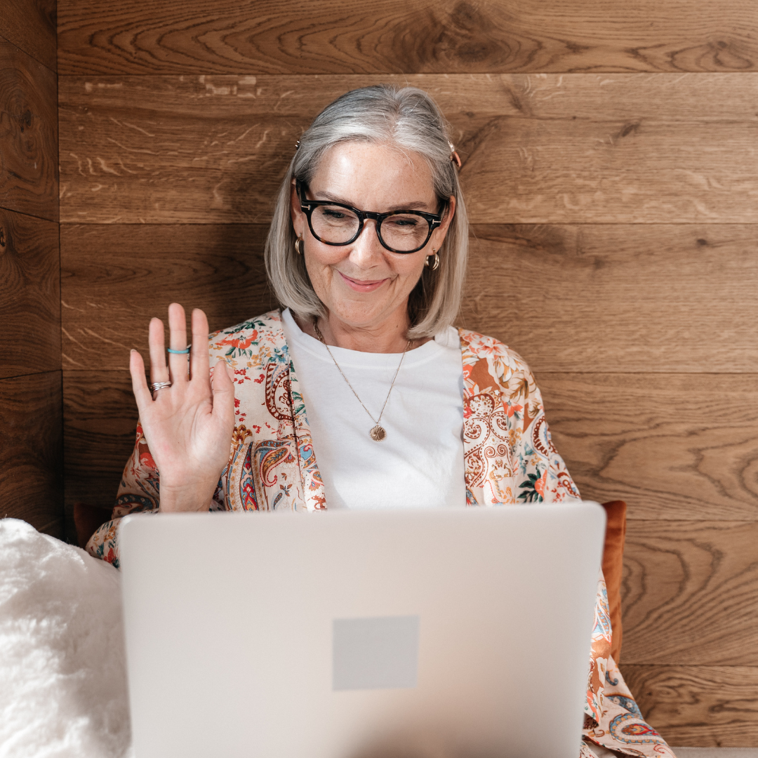 Older woman with gray hair, glasses, and earrings waving while sitting in front of a laptop against a wooden wall background.