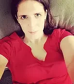 A woman with dark hair and fair skin wearing a red shirt, lying down on a gray surface, looking at the camera.