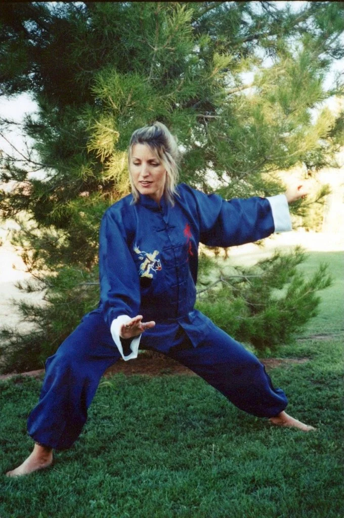 Woman practicing martial arts outdoors in traditional blue martial arts uniform with dragon embroidery, standing on grass in front of a pine tree, in a martial stance.