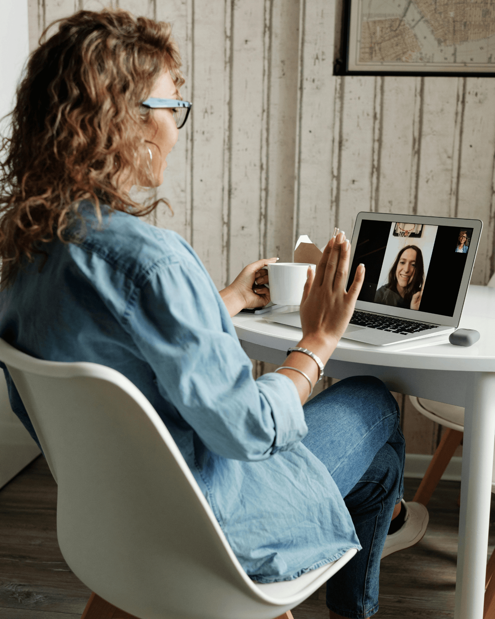 Woman with curly hair, glasses, and a blue shirt engages in a video call on her laptop, sitting at a white desk with a coffee mug, in a room with white wooden walls.
