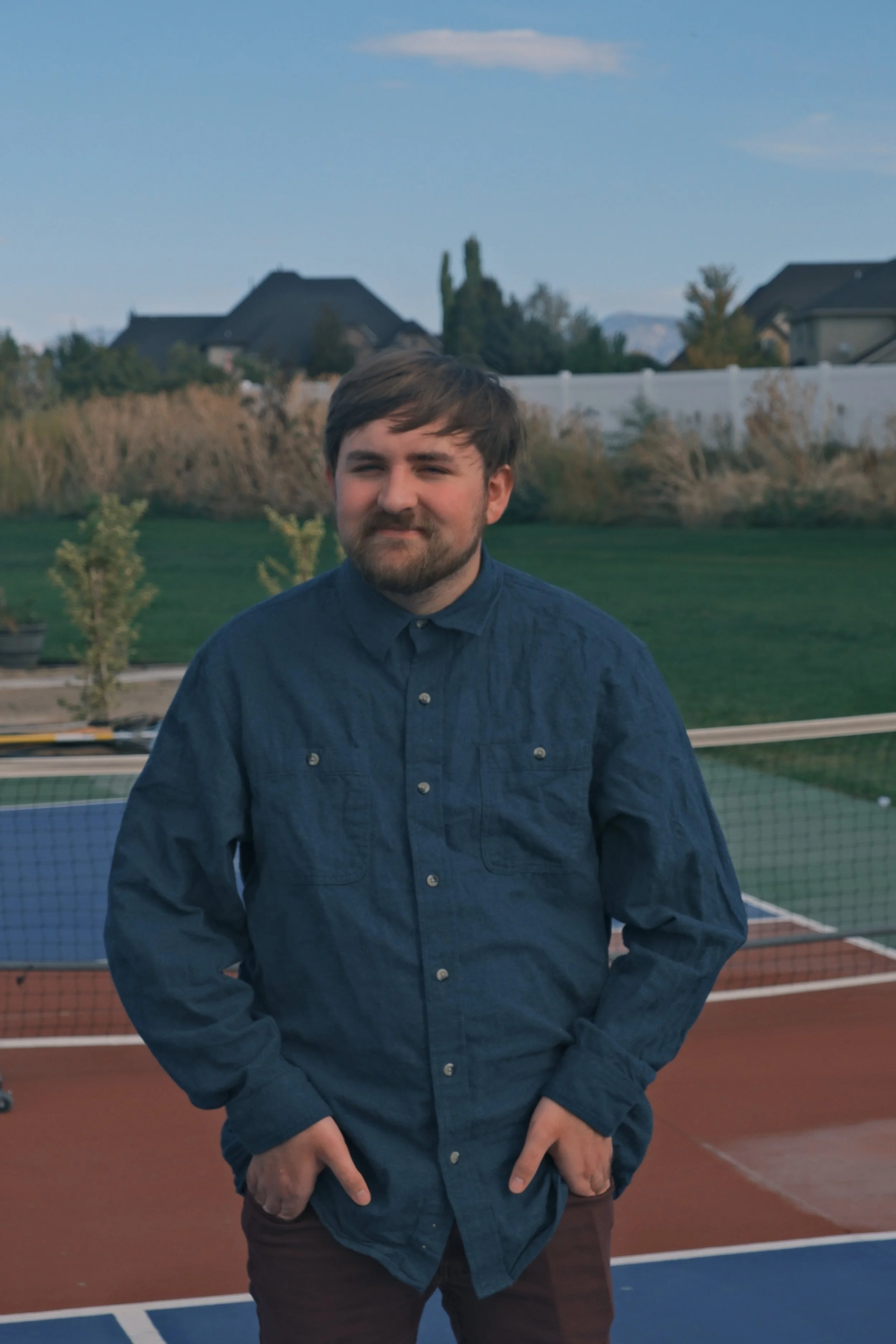 A young man with brown hair and a beard standing outdoors on a tennis court, wearing a blue button-up shirt and brown pants, with houses and trees in the background.