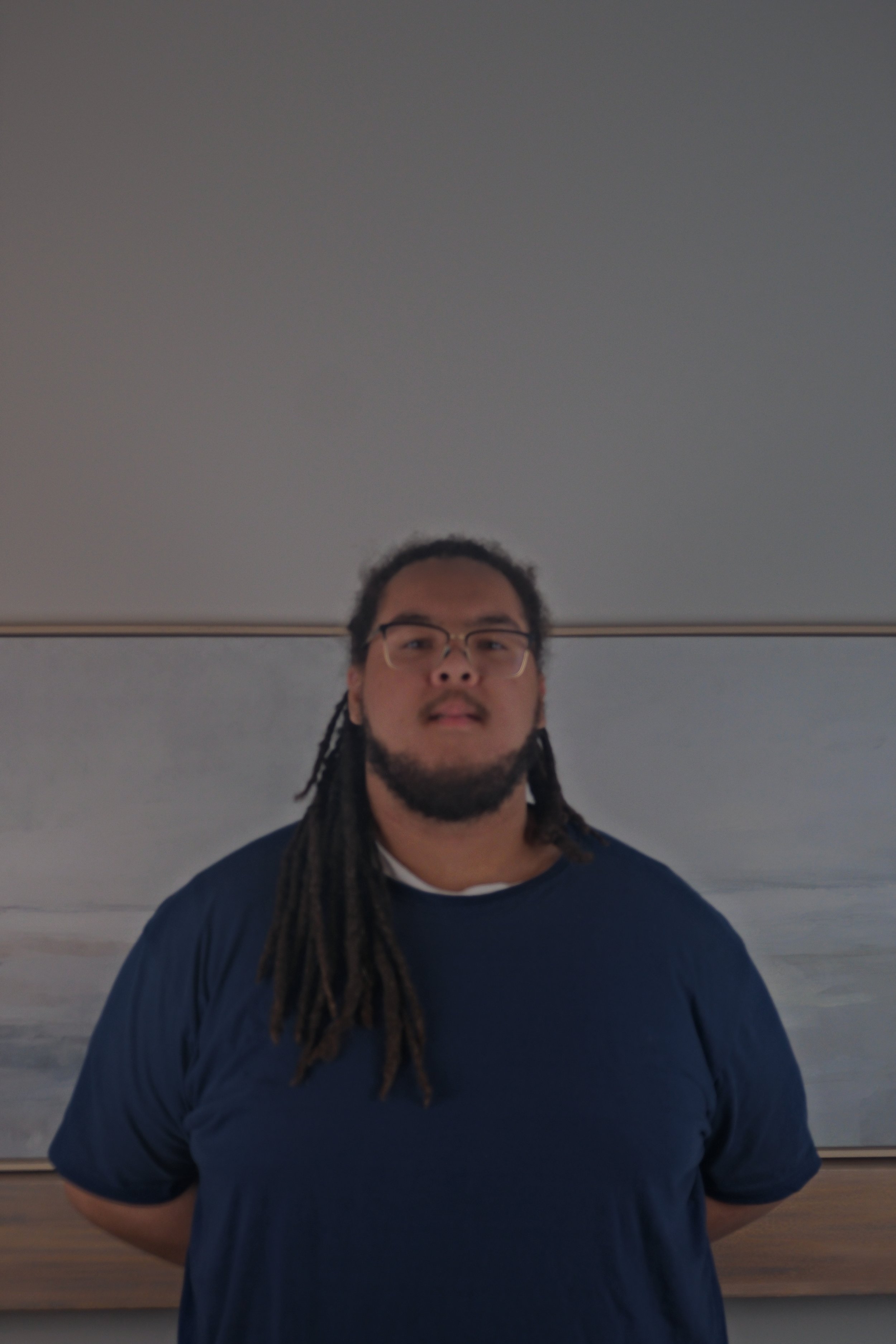 A man with glasses and long dreadlocks standing indoors in front of a plain wall with a framed landscape painting.