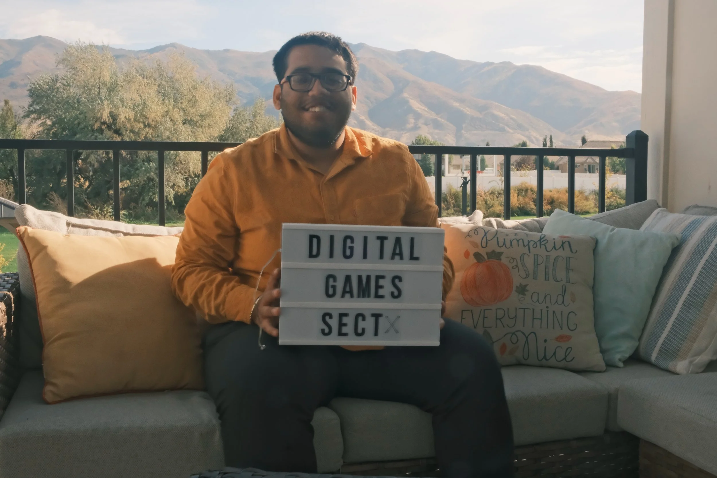 A man wearing glasses and a mustard-colored shirt sits on a sofa outdoors, holding a lightbox sign that reads 'DIGITAL GAMES SECT x'. Behind him, mountains and trees are visible, along with a decorative pillow that says 'Pumpkin spice and everything nice' and another striped pillow.