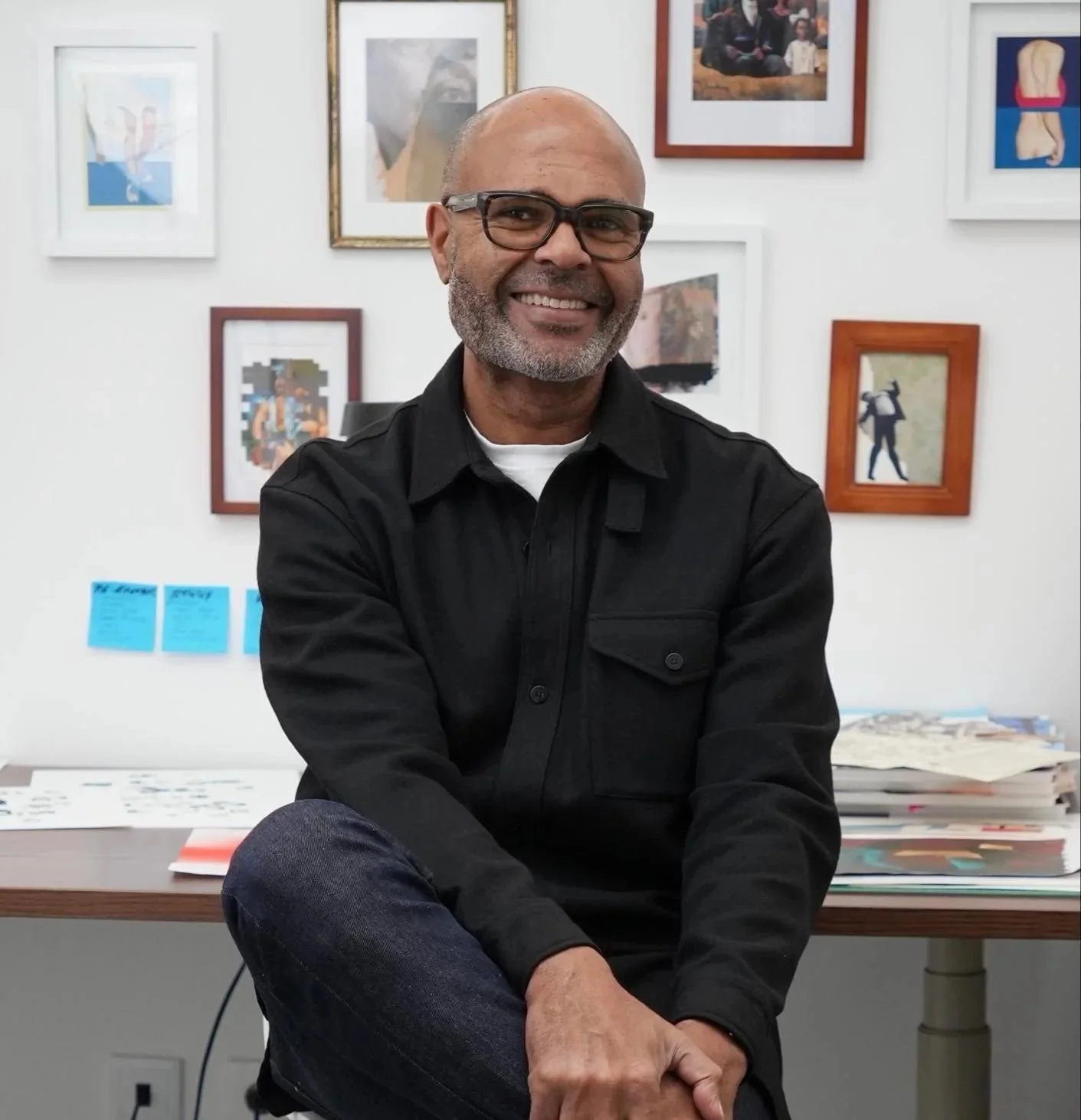 A smiling middle-aged man with glasses and a gray beard sitting with crossed legs in an art gallery or office, with framed artwork on the white wall behind him and stacked papers on a desk.