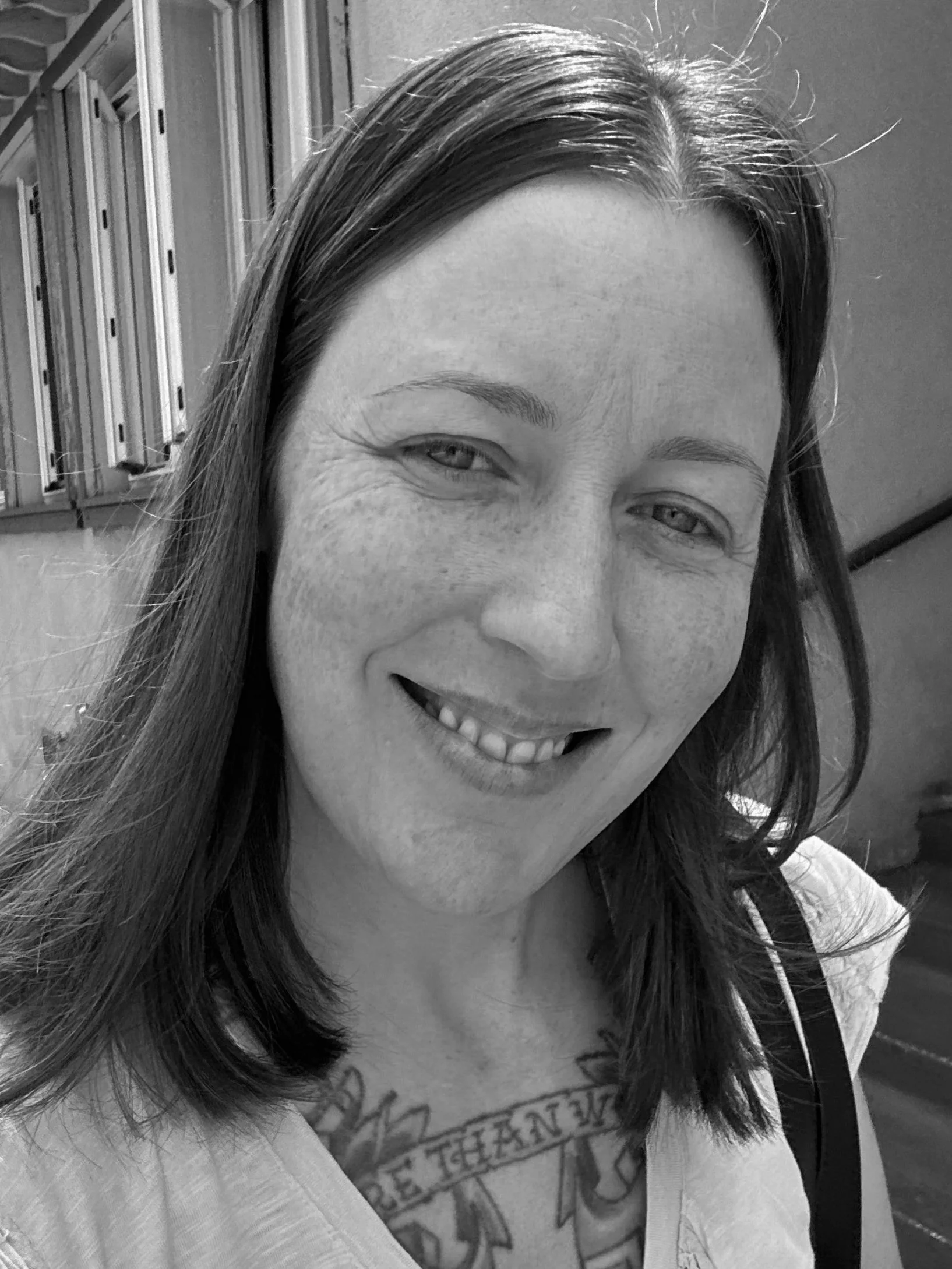 Black and white close-up photo of a smiling woman with long hair, visible tattoo on her chest, indoors with lockers in the background.