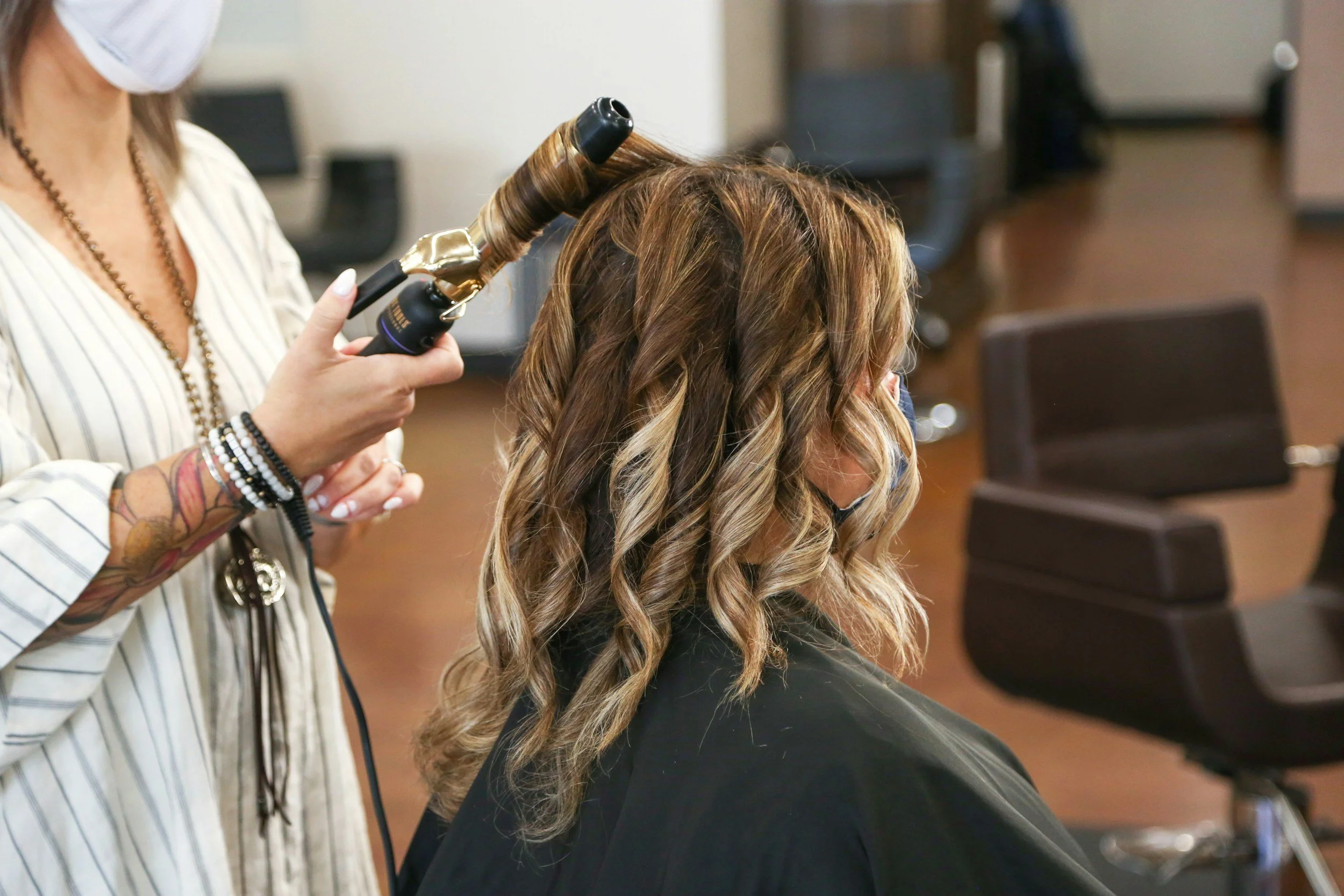 A hairstylist curling a woman's hair using a curling iron in a salon, both wearing masks.