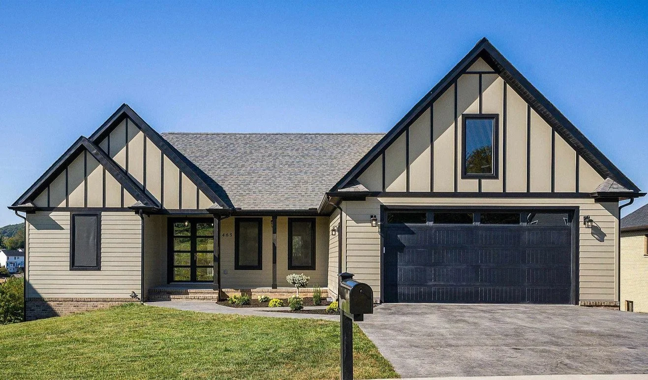 Front view of a modern house with a gray roof, beige siding with black trim, a large black garage door, and a front porch with steps.