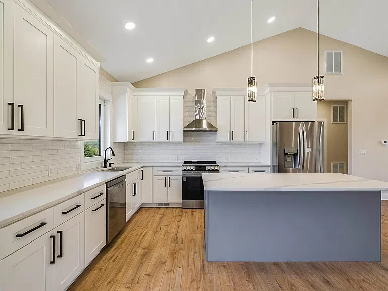 Modern kitchen with white cabinets, stainless steel appliances, a large kitchen island with light blue base, pendant lighting, hardwood floors, and a vaulted ceiling.