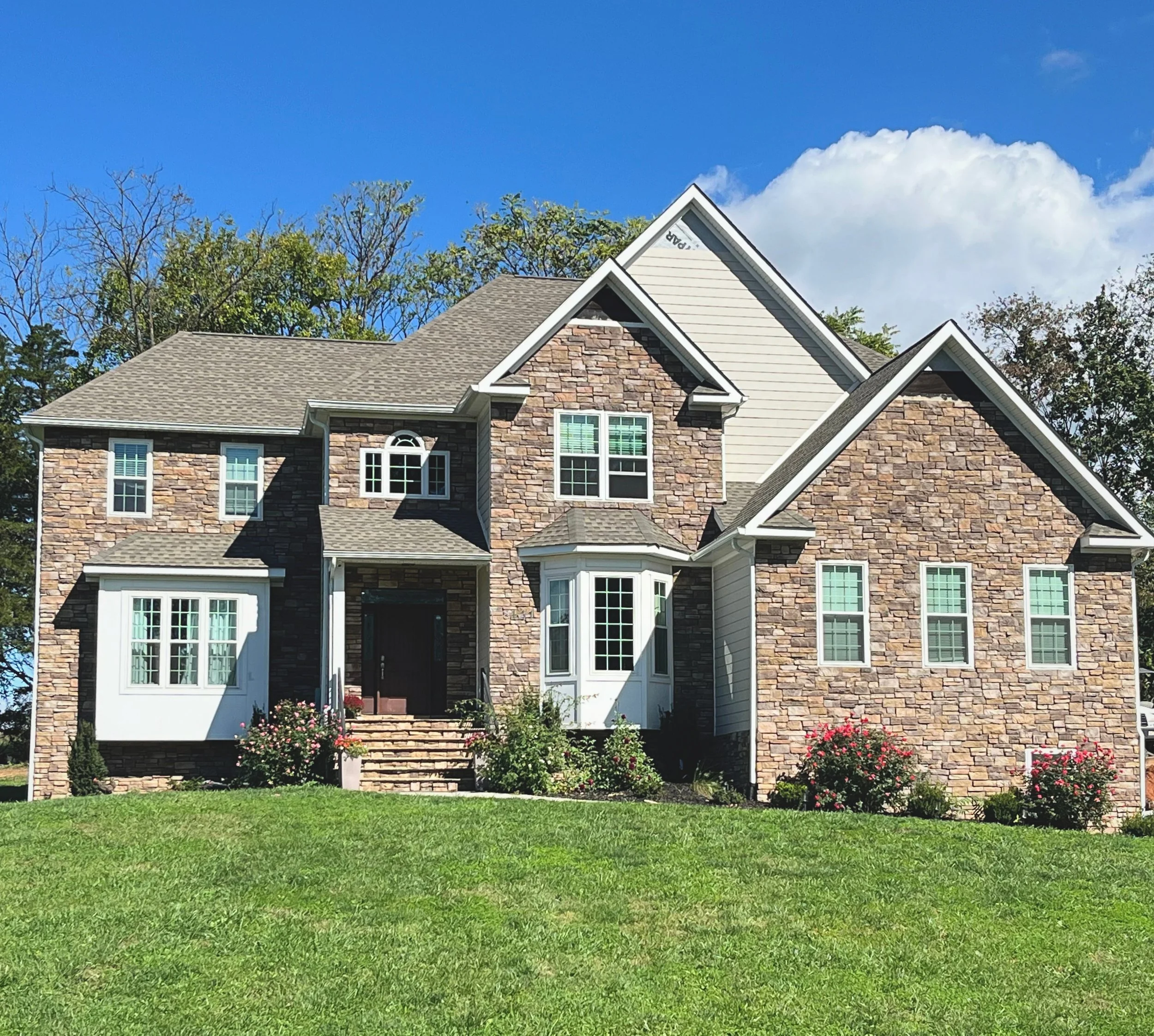A two-story house with brick and beige siding exterior, multiple gables, and white-framed windows, surrounded by a green lawn with bushes and flowers.