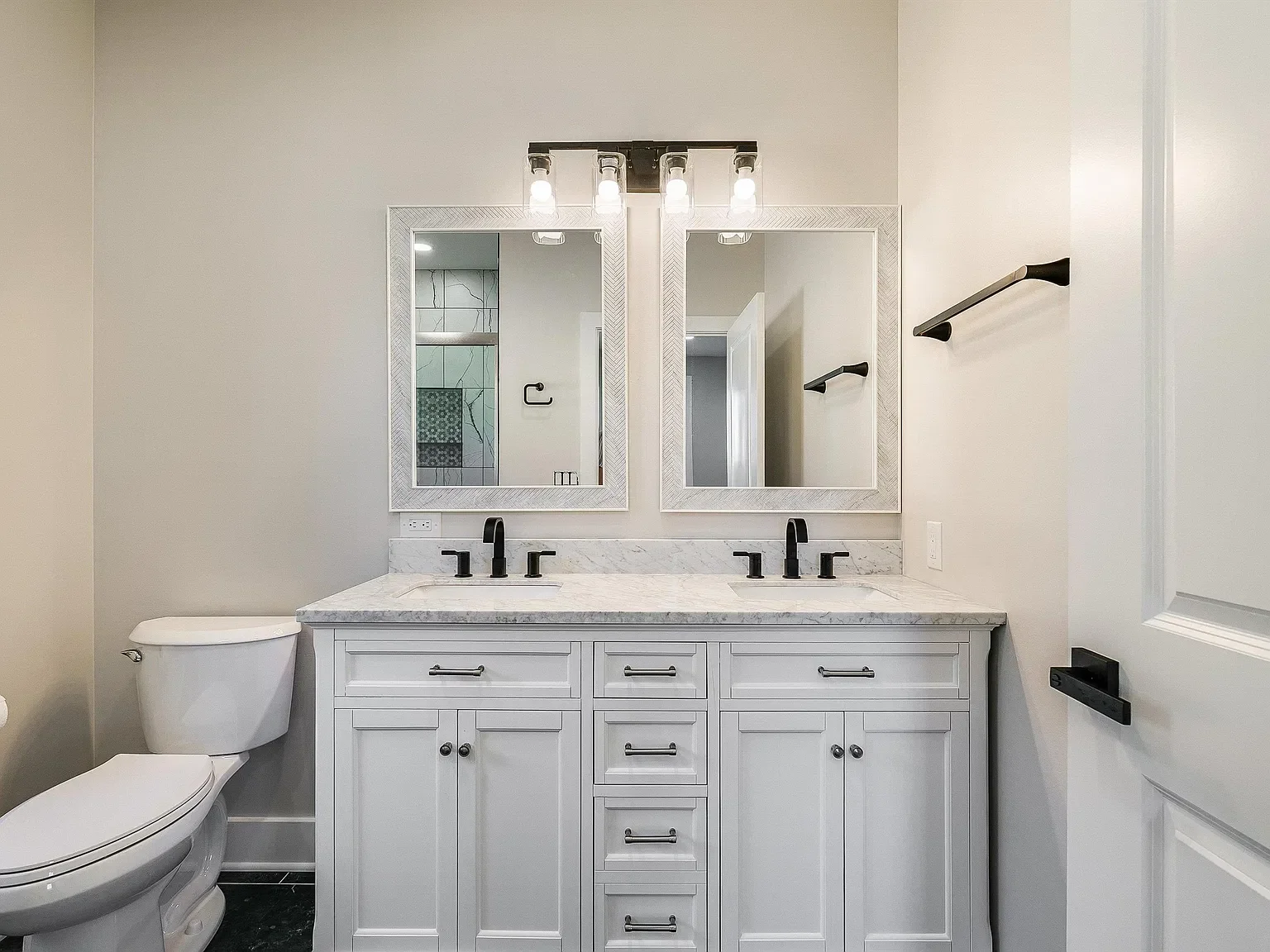 Modern bathroom with white vanity, marble countertop, and two framed mirrors. Two black faucets and handles, black towel bars, and a toilet on the left.