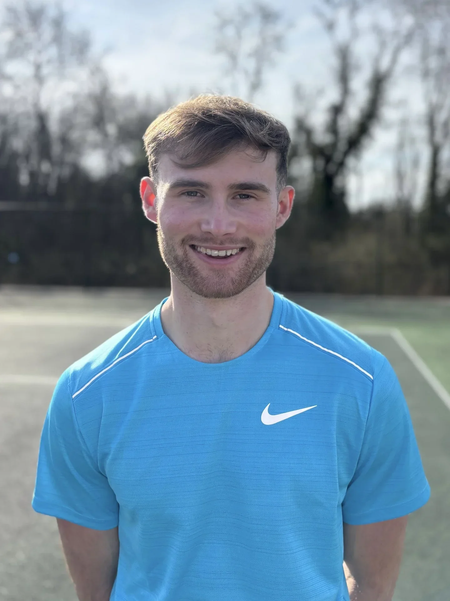 Young man smiling, wearing a blue Nike athletic shirt, standing outdoors on a sports field, with trees and a cloudy sky in the background.