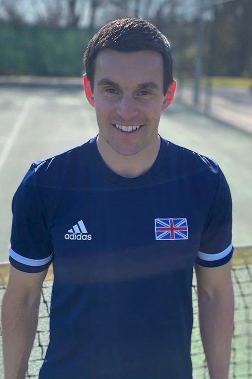 A young man with dark hair smiling while standing outdoors on a tennis court wearing a navy blue Adidas sports jersey with a British flag emblem.