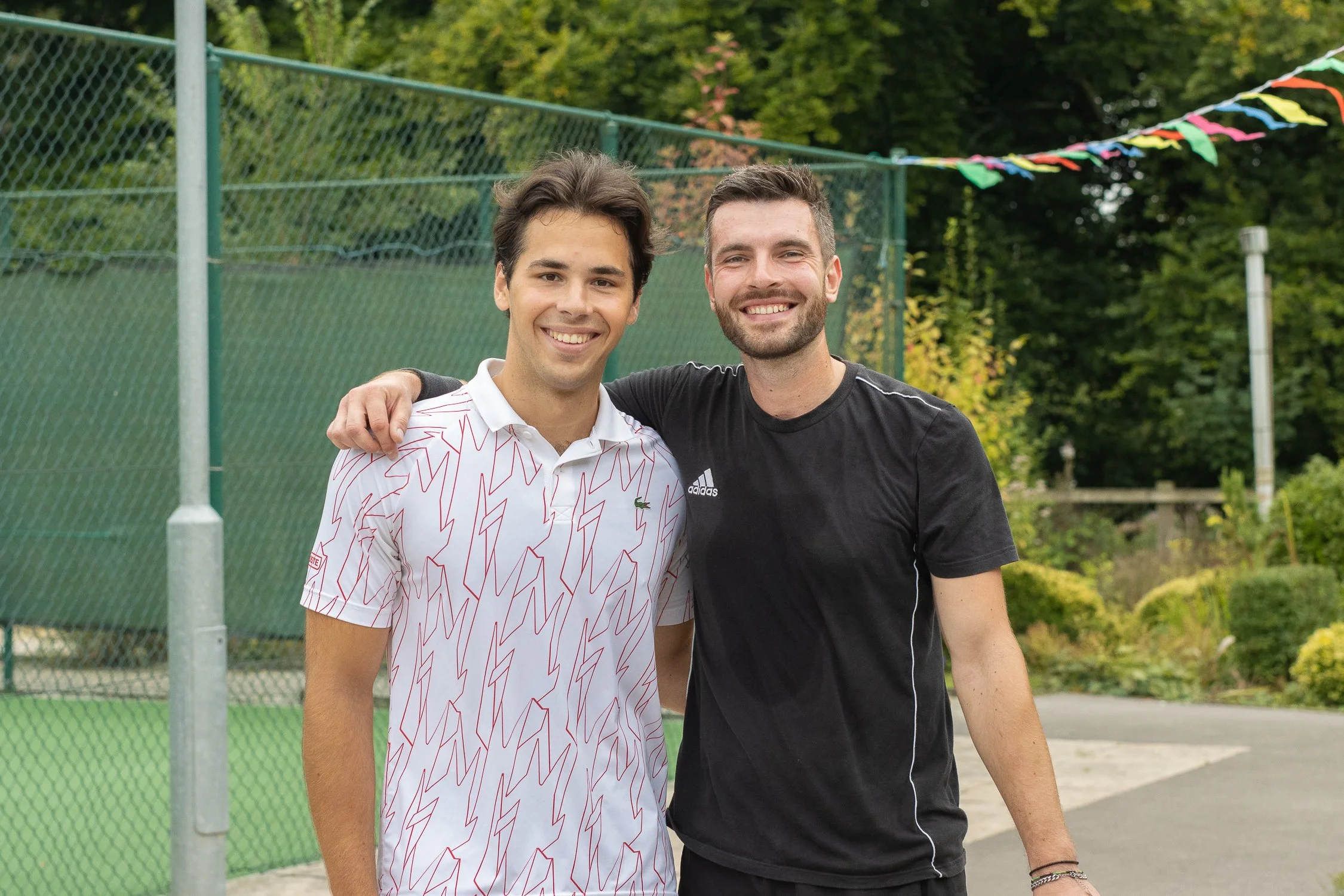 Two smiling men standing outdoors near a tennis court with a green fence and colorful pennant banners in the background.
