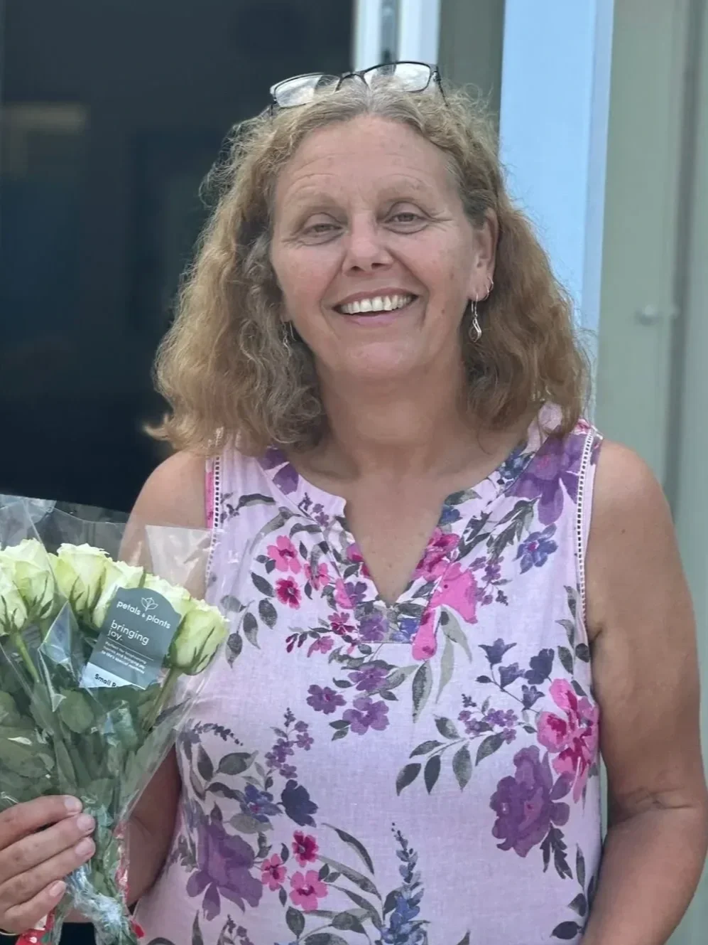 Smiling woman with curly hair holding a bouquet of white roses, wearing a sleeveless floral top, glasses on her head, and earrings, standing in front of a blue and black background.