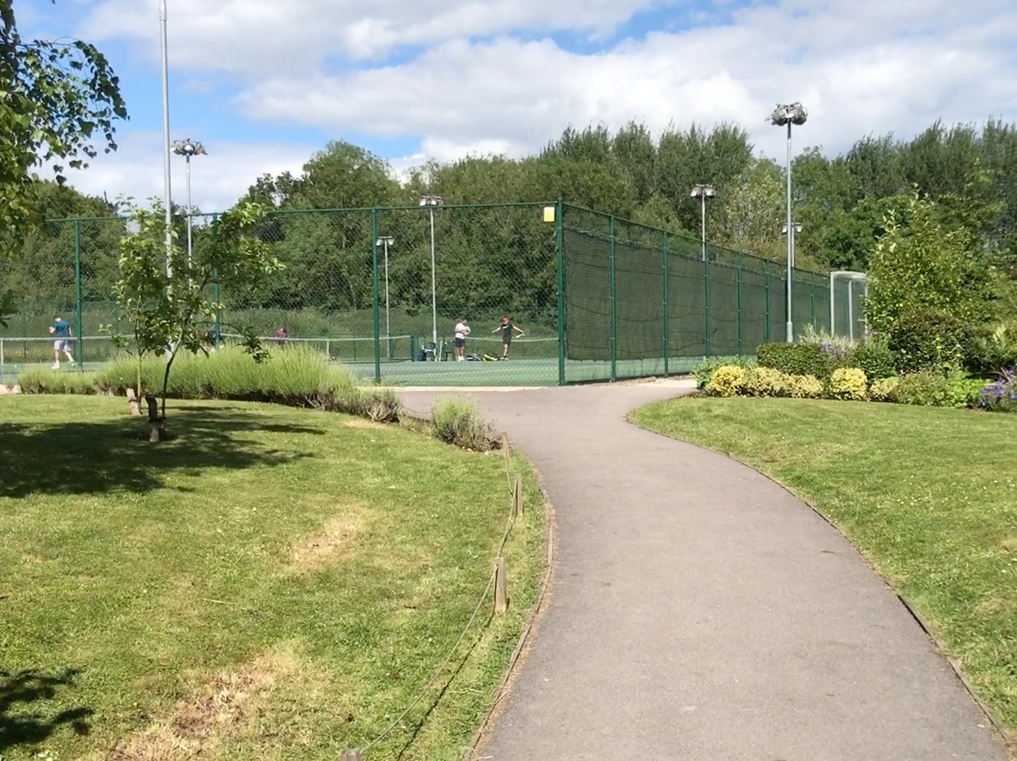 A pathway in a park leading to a fenced tennis court with players, surrounded by green grass, trees, and bushes under a partly cloudy sky.