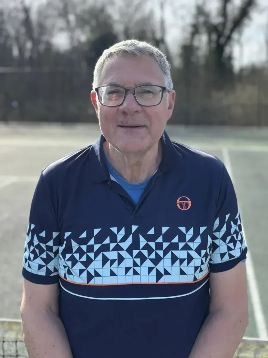 A middle-aged man with glasses, short gray hair, and light skin standing outdoors on a tennis court, wearing a navy blue sports shirt with a geometric pattern and a small logo.