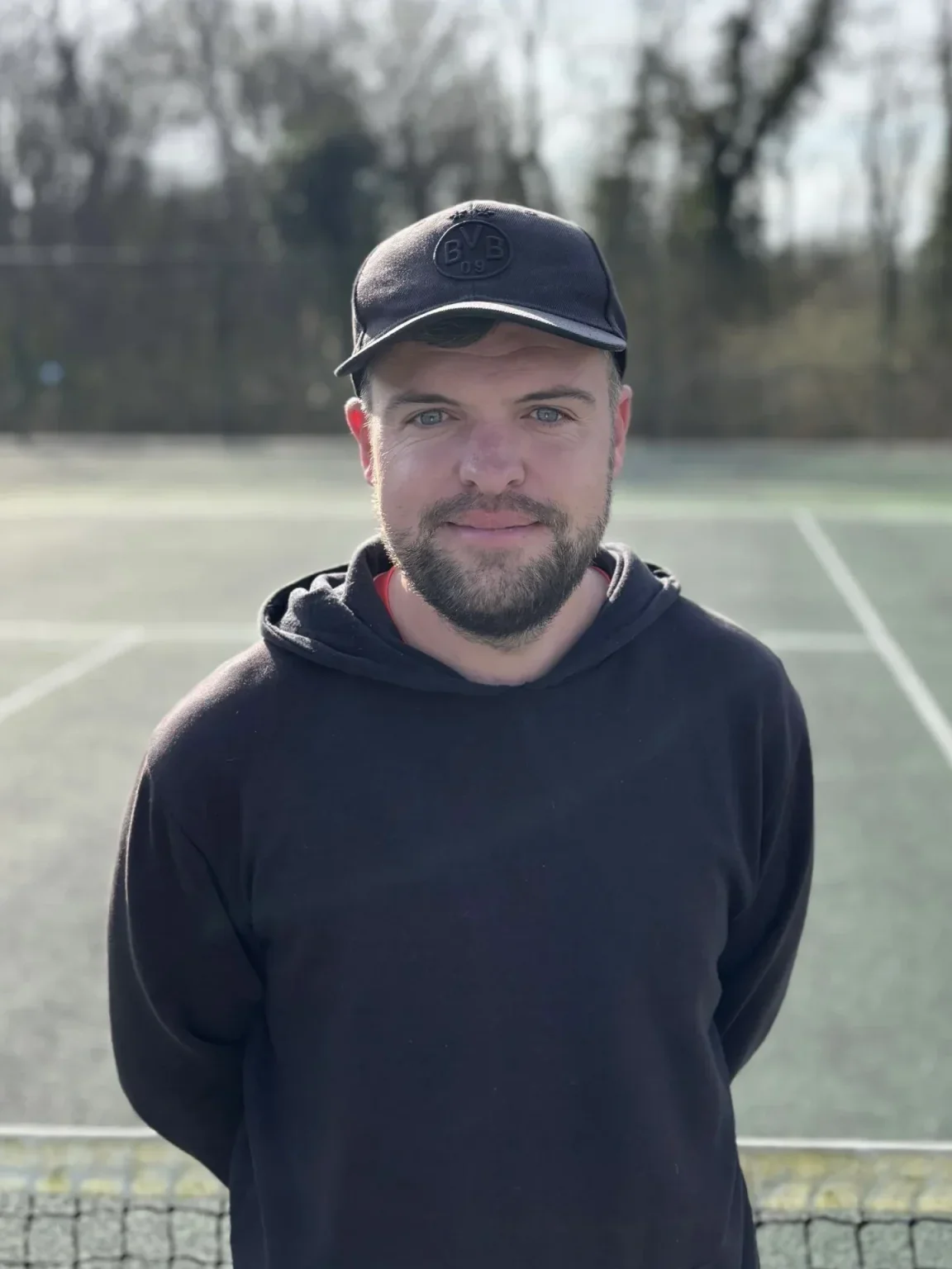 A young man with a beard and blue eyes standing on a tennis court during daytime, wearing a black hoodie and a black cap.