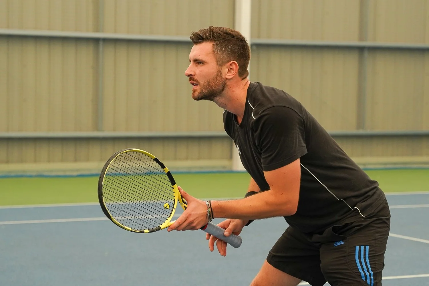 A man playing tennis on an indoor court, holding a tennis racket, and preparing to hit the ball.
