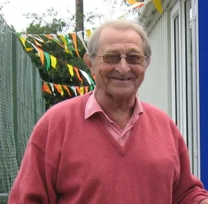 An elderly man with glasses and gray hair standing outdoors, smiling, wearing a pink sweater over a collared shirt.