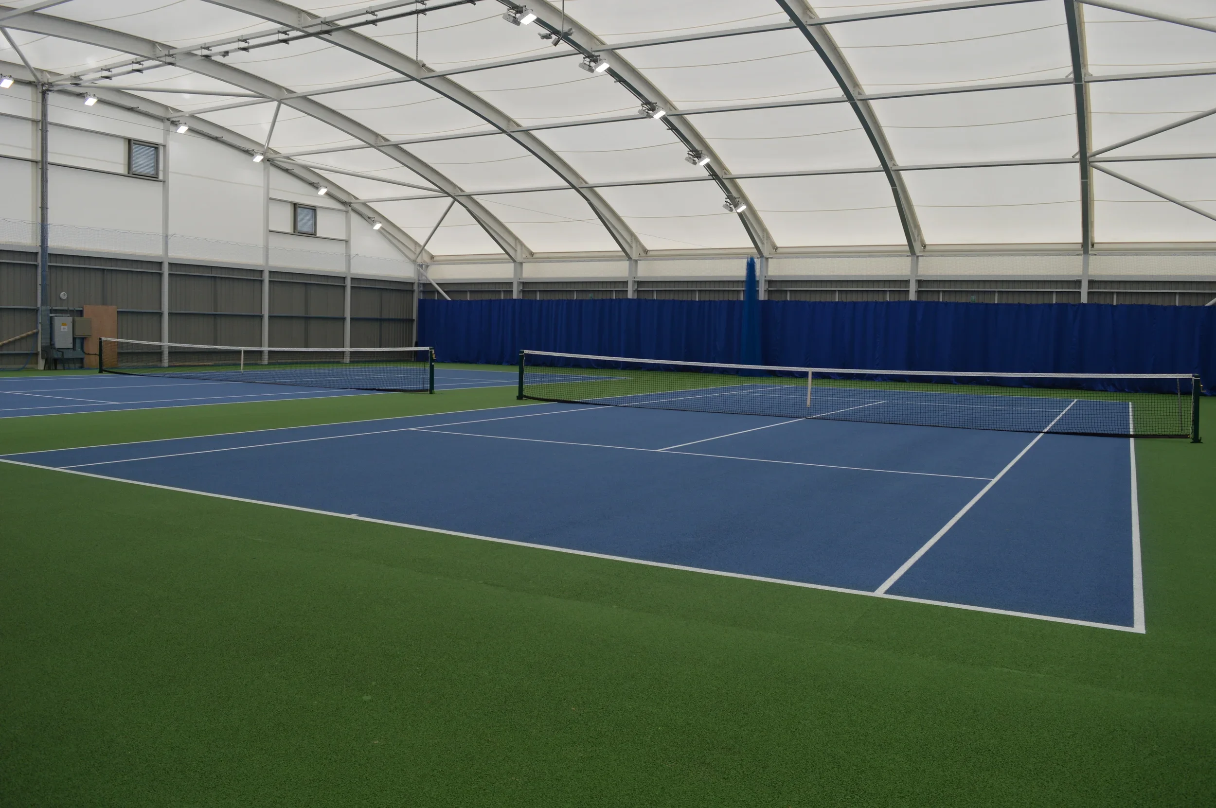 Indoor tennis courts with blue surfaces, netting, and a high, arched ceiling.
