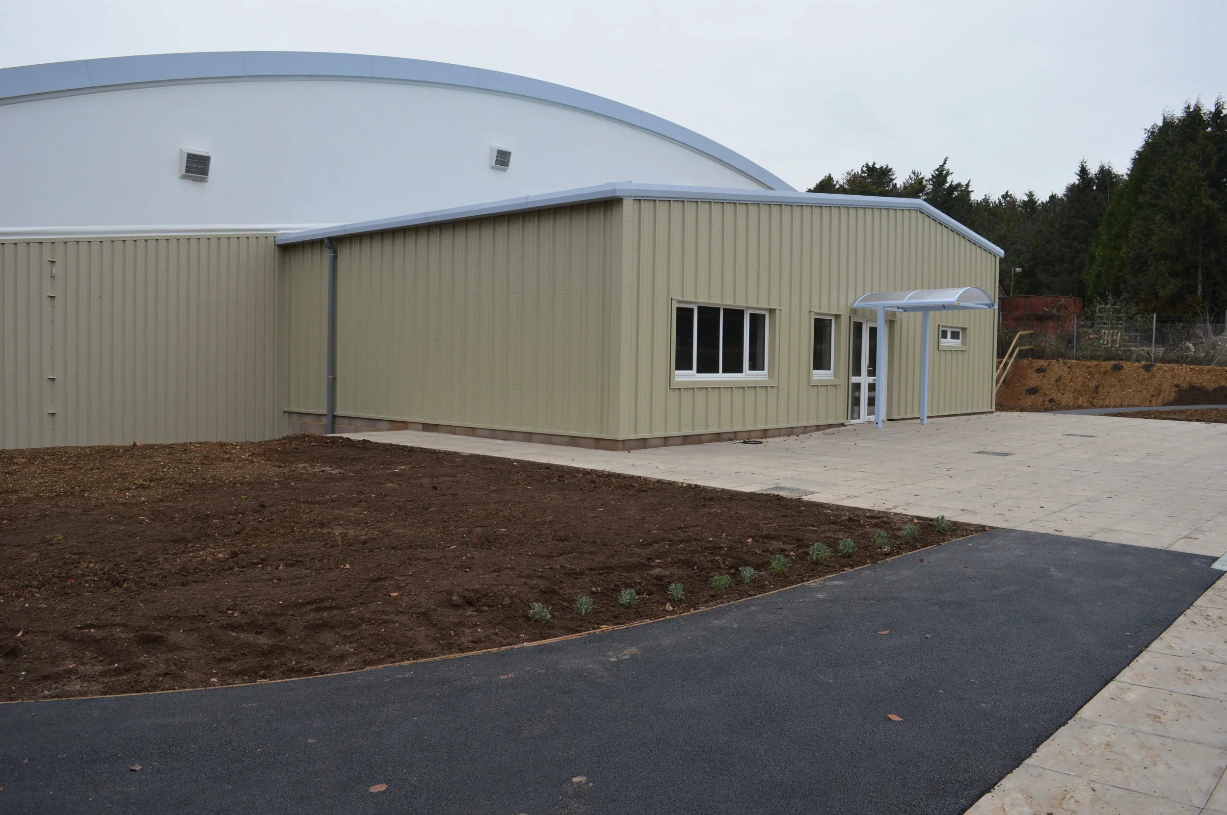 A modern building with beige metal siding and multiple windows, a small covered entryway, and an adjacent paved area, with a garden bed and a new paved pathway in the foreground.