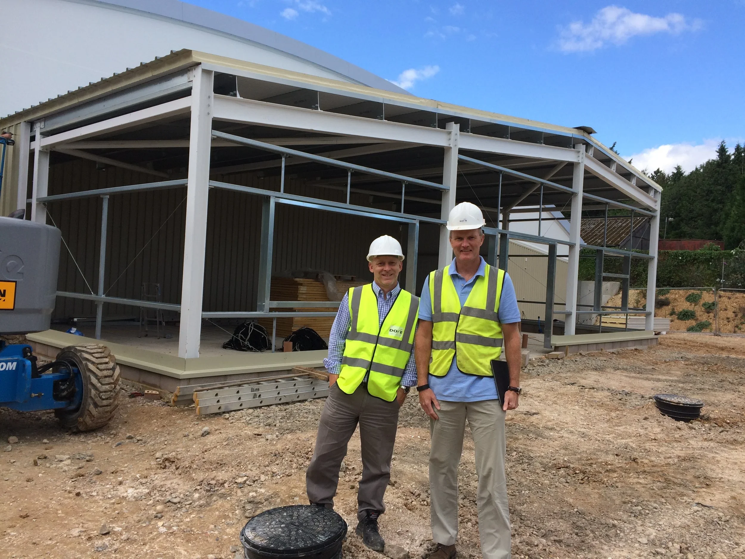 Two men wearing white hard hats and yellow safety vests standing in front of a construction site with a partially built structure and construction equipment.