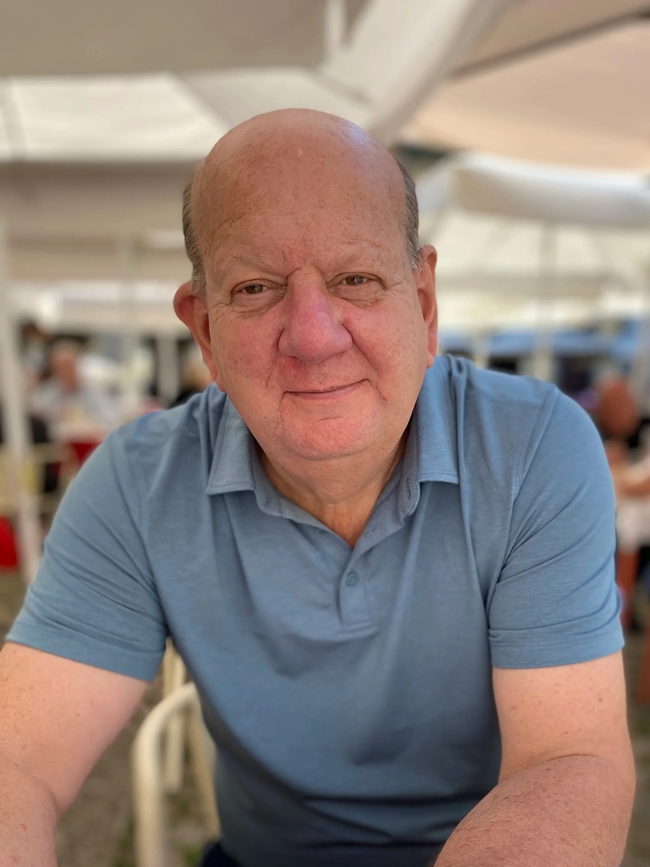 A close-up of an older man with a bald head and wearing a blue polo shirt, sitting outdoors under a white canopy at a restaurant or café, with a blurred background of other diners.