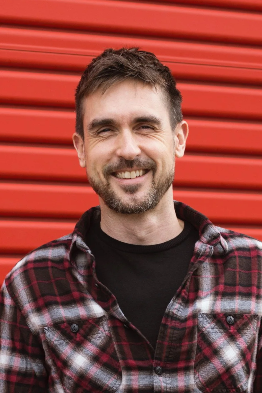 A smiling man with dark hair and a beard, wearing a black shirt and a red and black plaid flannel, standing outdoors in front of a red corrugated metal wall.