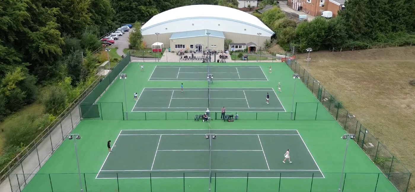 Two tennis courts with players, a building in the background, surrounded by trees and parking lot.